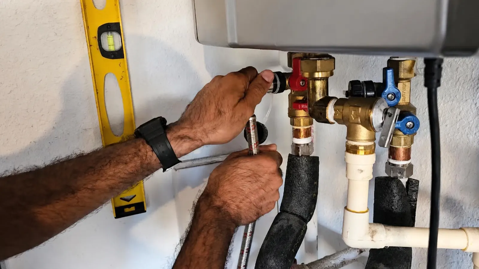 Technician connecting isolation valves and insulated pipes beneath a wall-mounted tankless water heater, with a level held against the wall